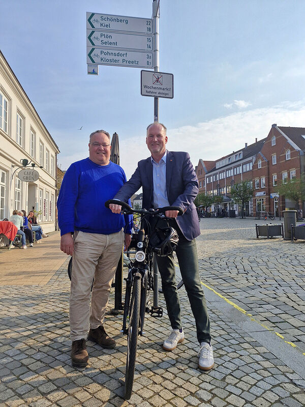 Bild vergr&ouml;&szlig;ern: Auf dem Foto sind Lutz Schlünsen, Vorsitzender des Ausschusses für Wirtschaft, Energie und Tourismus und Landrat Björn Demmin mit Fahrrad zu sehen. Beide stehen unter einem neuen Radwegweiser in der Fußgängerzone der Stadt Plön.