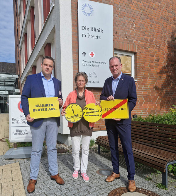 Bild vergr&ouml;&szlig;ern: Auf dem Foto sind von links nach rechts der Geschäftsführer der Klinik Preetz Sven Sandberg, Kreispräsidentin Hildegard Mersmann und Landrat Björn Demmin zu sehen. Sie halten zur Verdeutlichung der Problematik verschiedene Schilder vor sich. "Kliniken bluten aus", eine Uhr deren Zeiger auf kurz vor 12 stehen, "diese Einrichtung ist in Gefahr" und ein Schild im Stil eines Ortsausgangsschildes, auf dem der Schriftzug Krankenhaus steht, der mit einem roten diagonalen Balken durchgestrichen ist.