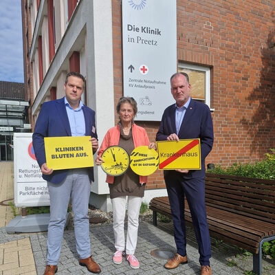 Auf dem Foto sind von links nach rechts der Geschäftsführer der Klinik Preetz Sven Sandberg, Kreispräsidentin Hildegard Mersmann und Landrat Björn Demmin zu sehen. Sie halten zur Verdeutlichung der Problematik verschiedene Schilder vor sich. "Kliniken bluten aus", eine Uhr deren Zeiger auf kurz vor 12 stehen, "diese Einrichtung ist in Gefahr" und ein Schild im Stil eines Ortsausgangsschildes, auf dem der Schriftzug Krankenhaus steht, der mit einem roten diagonalen Balken durchgestrichen ist.