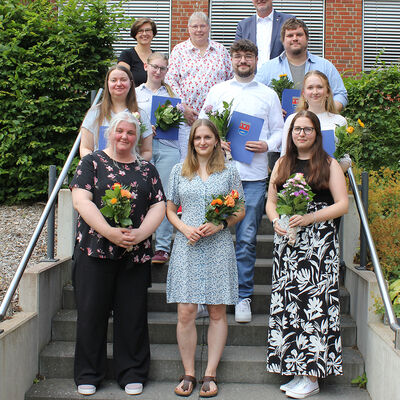 Auf dem Foto sind die diesjährigen Absolventinnen und Absolventen der Kreisverwaltung Plön zu sehen. Sie stehen mit ihren Urkunden und kleinen Blumensträußen zusammen mit der Gleichstellungsbeauftragten Yvonne Deerberg, der Personalratsvorsitzenden Christel Pfau und Landrat Björn Demmin auf dem Treppenaufgang zum Kreistagssitzungssaal.